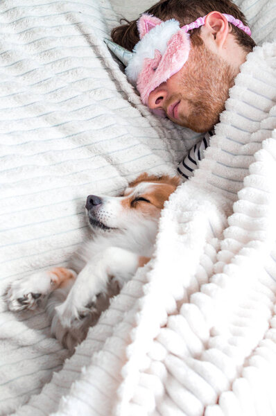 Closeup portrait of a young man in a pink mask sleeping in a bed under a rug with his dog. Vertical photo