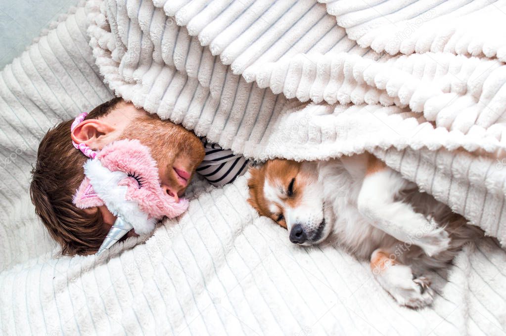 Closeup portrait of a young man in a pink mask sleeping in a bed under a rug with his dog