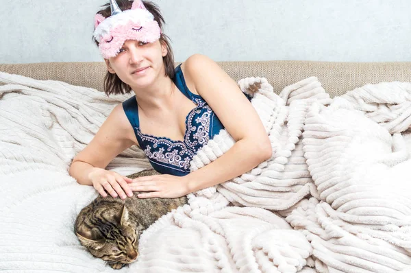 Retrato de uma jovem de manhã com uma máscara de dormir no rosto e pijama. Perto está um gato cinzento dormindo. Manhã de conceito. Mulher é feliz e sorrindo — Fotografia de Stock