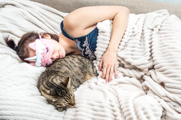 Retrato de uma jovem mulher com uma máscara de sono no rosto e pijama adormecido abraçando seu gato. Conceito de descanso e sono . — Fotografia de Stock