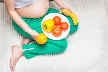 Beyaz bir tabak yemekle yatakta oturan hamile bir kadın. Meyve ve sebzeler. Hamile kadın beslenme kavramı