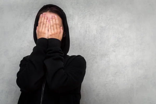 young woman in a black sweater and a hood on her head covers her face with her hands. The concept of mental health. Depression. Stress. Negative emotion.