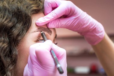 Kaş ustası, kaş kalemi olan genç bir kadının kaşlarının şeklini çizer. Güzellik salonunda kınalı kaş boyama prosedürü