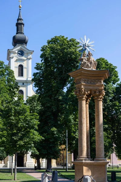 Saati olan tarihi bir kilise kulesinin manzarası, yanında parlak mavi gökyüzünün altında yemyeşil ağaçlarla çevrili, ayrıntılı bir heykel var..