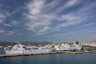 SPLIT, CROATIA, 08.15.2020: Ferry docked in a coastal town's harbor, surrounded by lush green hills under a clear blue sky.