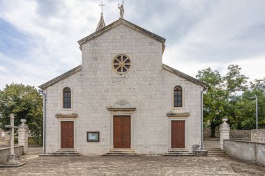 Çan kulesi ve haçı olan yıpranmış taş bir kilise sağlam bir taş duvarın yanında duruyor. Yeşil kapılar ve taşralı duvarcılık zamansız kırsal mimariyi ve sessiz inancı çağrıştırıyor.
