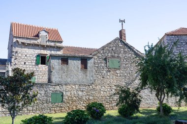 Ağaçlarla ve manikürlü bahçeyle çevrili yeşil panjurlu tarihi bir taş bina. Parlak gün ışığı desenli duvarlar, çatı kiremitleri ve sakin, kırsal bir kırsal ortam vurguluyor..