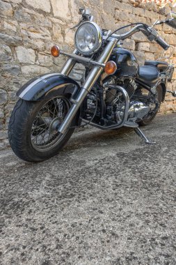 Klasik bir siyah kruvazör motosikleti, krom aksanlı ve klasik stili olan taştan bir duvarın yanına park edilmiş. Seyahat, yaşam tarzı ya da otomotiv temaları için basit, engebeli ve zamansız