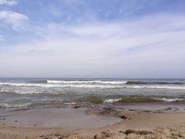 Dalgalar Litvanya 'nın Palanga kentindeki Baltık Denizi' nin kumlu sahillerine çarpar. Gökyüzü parçalı bulutludur, mavi ve beyaz renklerin bir karışımını yaratır. Koyu, ıslak kum ve dağınık kayalar görülebilir.