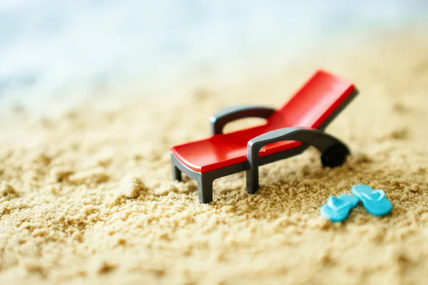 red toy deck chair and small blue flip-flops on the sand next to the sea in summer