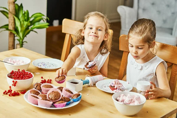 Duas belas irmãs em vestidos brancos decorar e comer deliciosos cupcakes — Fotografia de Stock