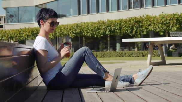 Mujer en una camiseta blanca sentada frente a un edificio de oficinas y bebiendo jugo de naranja — Vídeos de Stock