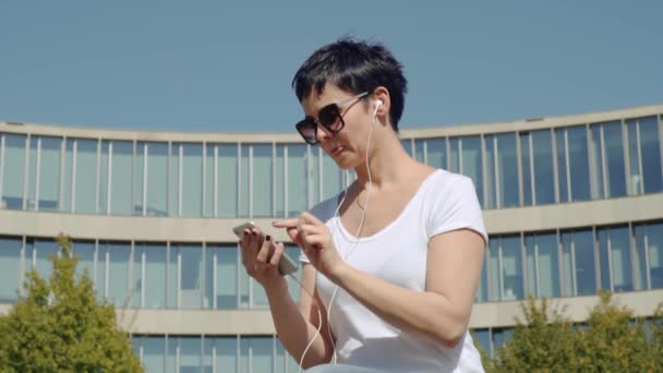 Business kvinna i en vit T-shirt sitter framför en kontorsbyggnad och lyssnar på musik — Stockvideo