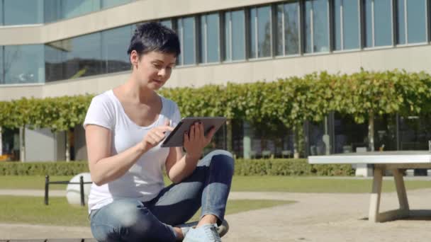 Mulher em uma camiseta branca sentada na frente de um prédio de escritórios e trabalhando em um tablet — Vídeo de Stock