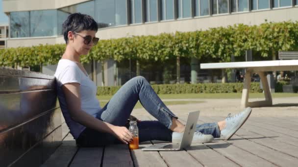 Женщина в белой футболке сидит перед офисным зданием и пьет апельсиновый сок — стоковое видео