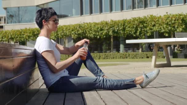 Woman in a white T-shirt sitting in front of an office building and drinking orange juice — Stock Video