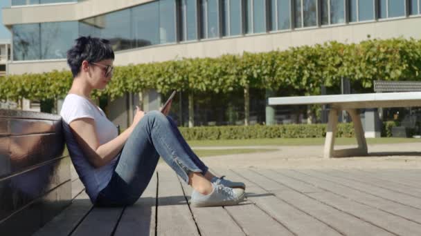 Женщина в белой футболке сидит перед офисным зданием и работает на планшете — стоковое видео