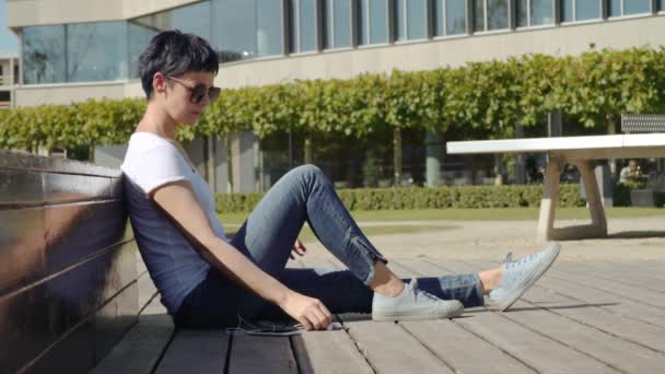 Business woman in a white T-shirt sits in front of an office building and listens to music — Stock Video
