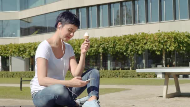 Frau mit Eiscreme sitzt vor einem Bürogebäude und hört Musik am Telefon — Stockvideo