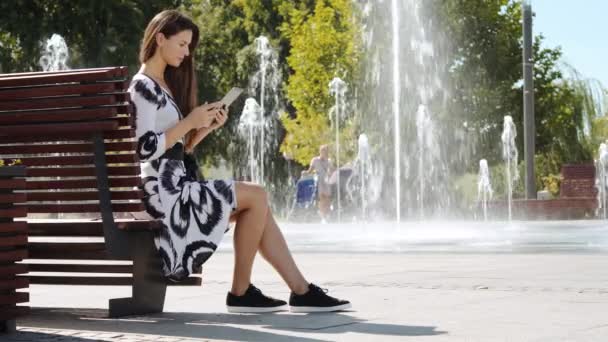 Młoda kobieta w sukni na ławce i działa na tablecie — Wideo stockowe