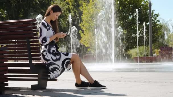 Jonge zakenvrouw in een jurk zittend op een bankje en praten over de telefoon — Stockvideo