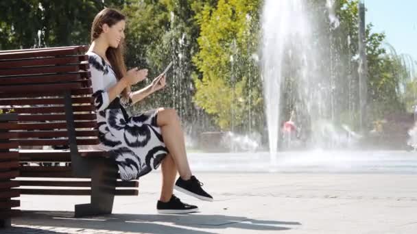 Joven mujer de negocios en un vestido sentado en un banco y hablando en la tableta — Vídeos de Stock