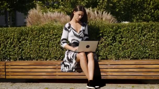 Jonge zakenvrouw in een jurk die zittend op een bankje met een telefoon in de hand en een computer — Stockvideo