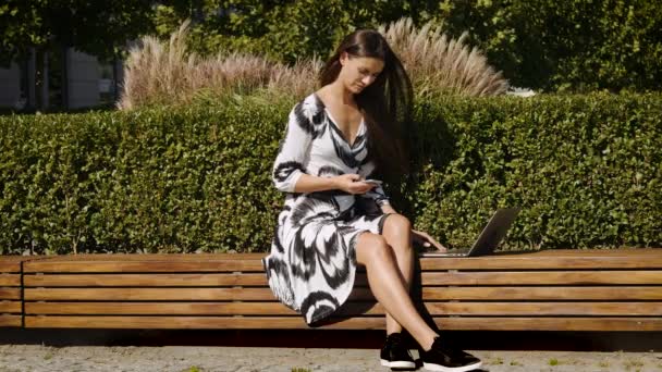 Tânără femeie de afaceri într-o rochie așezată pe o bancă cu un telefon în mână și un computer — Videoclip de stoc