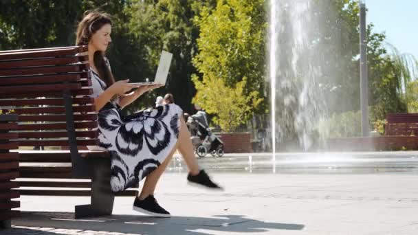 Νεαρή γυναίκα σε ένα φόρεμα που κάθεται σε ένα παγκάκι και λειτουργεί στον υπολογιστή — Αρχείο Βίντεο