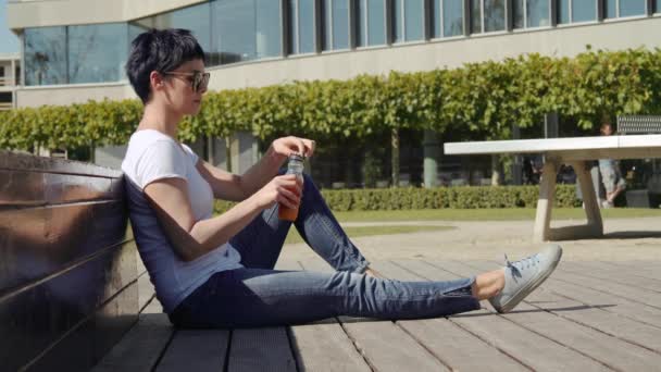 Frau im weißen T-Shirt sitzt vor einem Bürogebäude und trinkt Orangensaft — Stockvideo