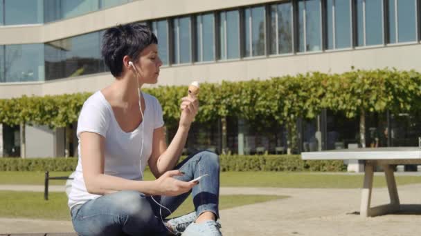 Vrouw met icecream zit tegenover een kantoorgebouw en luistert naar muziek op de telefoon — Stockvideo