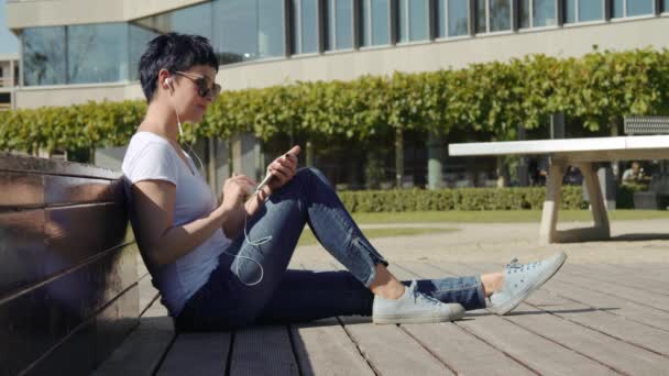 Mulher de negócios senta-se na frente de um prédio de escritórios e ouve música no telefone — Vídeo de Stock