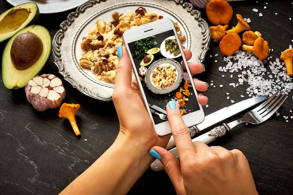 woman\'s hands are holding the phone and taking a picture of delicious scrambled eggs with mushrooms on a woody table