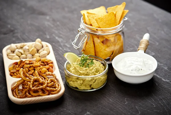 Bowl of guacamole jar with nachos and salty snacks on an old black wooden table — Stock Photo, Image