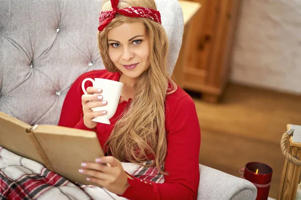 Junge schöne Frau liest ein Buch in einem Sessel mit Decke und Tee in der Weihnachtszeit — Stockfoto