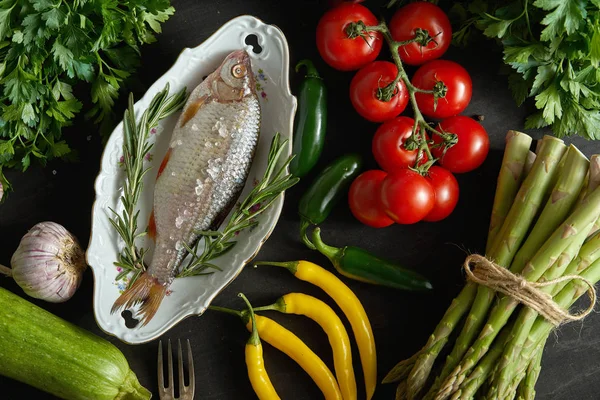 top view of one fresh raw dorada fish in a white dish with a set of vegetables on a black table