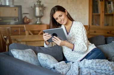 güzel genç gülümseyen kadın beyaz kazaklı bir tablet üzerinde gri bir kanepe ile oturan