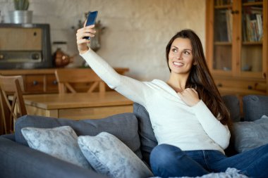 güzel genç gülümseyen kadın beyaz kazaklı selfie smartphone gri Divan taking
