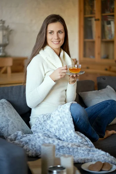 attractive young woman in sweater relaxes on a gray sofa at home and drinks tea with lemon