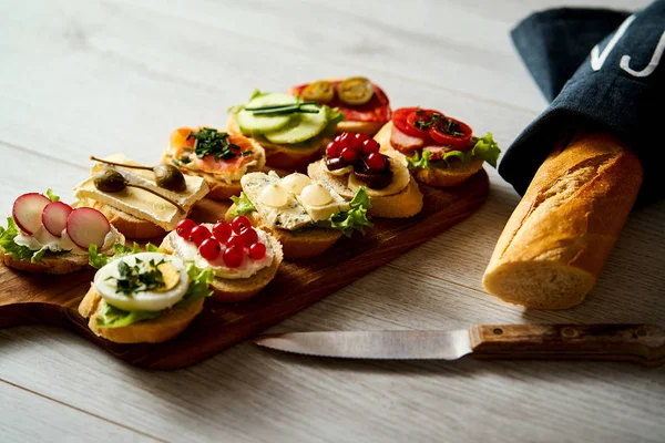 Wooden board with colorful mini sandwiches or tapas along with a knife and baguette on a table — Stock Photo, Image