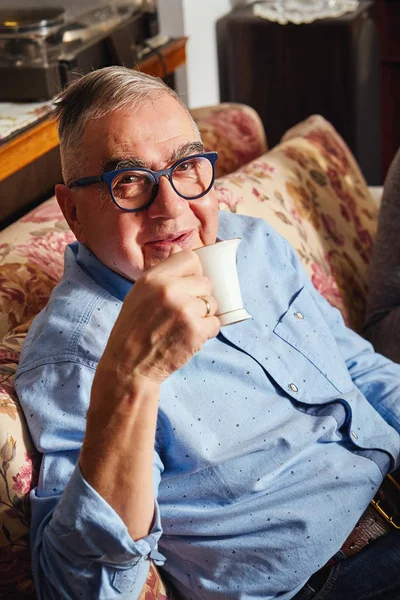Ευτυχισμένος ηλικιωμένους άνθρωπος κάθεται στον καναπέ και απολαμβάνει καφέ. — Φωτογραφία Αρχείου