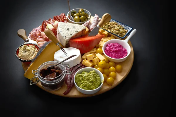 Placa antipasti com vários queijos e lanches de carne com húmus e azeitonas em tábua redonda de madeira — Fotografia de Stock