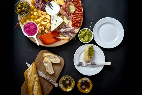 Antipasti board with cheese and meat snacks with white wine and baguette on board on black table — Stock Photo, Image