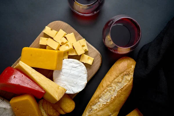 Wooden board with various types of cheese and baguette and two glasses of red wine on black table — Stock Photo, Image