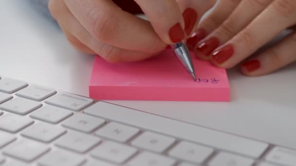 Primer plano de las manos femeninas escribir nota en el poste de color rosa junto al teclado de la computadora blanca — Vídeos de Stock