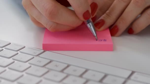 Закрывая женские руки, пишите записку на розовом столбе рядом с белой клавиатурой компьютера — стоковое видео