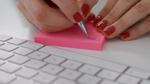 Primer plano de las manos femeninas escribir nota en el poste de color rosa junto al teclado de la computadora blanca — Vídeos de Stock