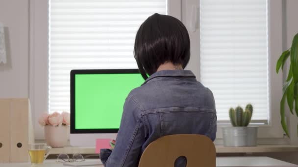 Вид ззаду жінки, що працює на клавіатурі комп'ютера і розмовляє по телефону перед дисплеєм з ізольованим зеленим екраном — стокове відео