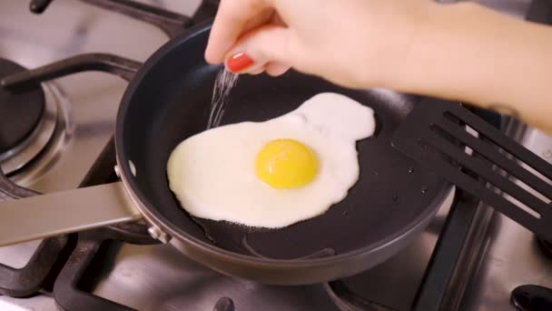 Vrouw in huis keuken frituren gebakken ei en het zout in de pan toe te voegen voor het ontbijt gaskachel — Stockvideo