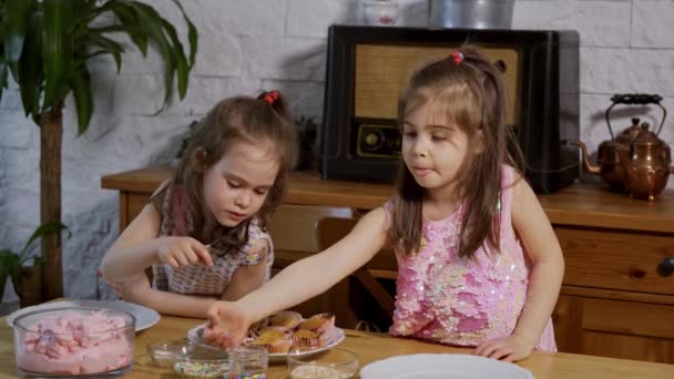 Due bambine si mettono una crema e decorano deliziosi cupcake su un tavolo di legno — Video Stock